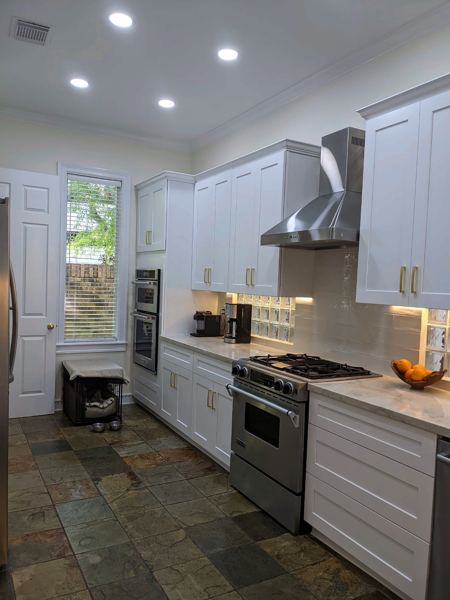 White shaker kitchen with gold pulls, stainless range hood, built-in double ovens, and natural slate tile flooring