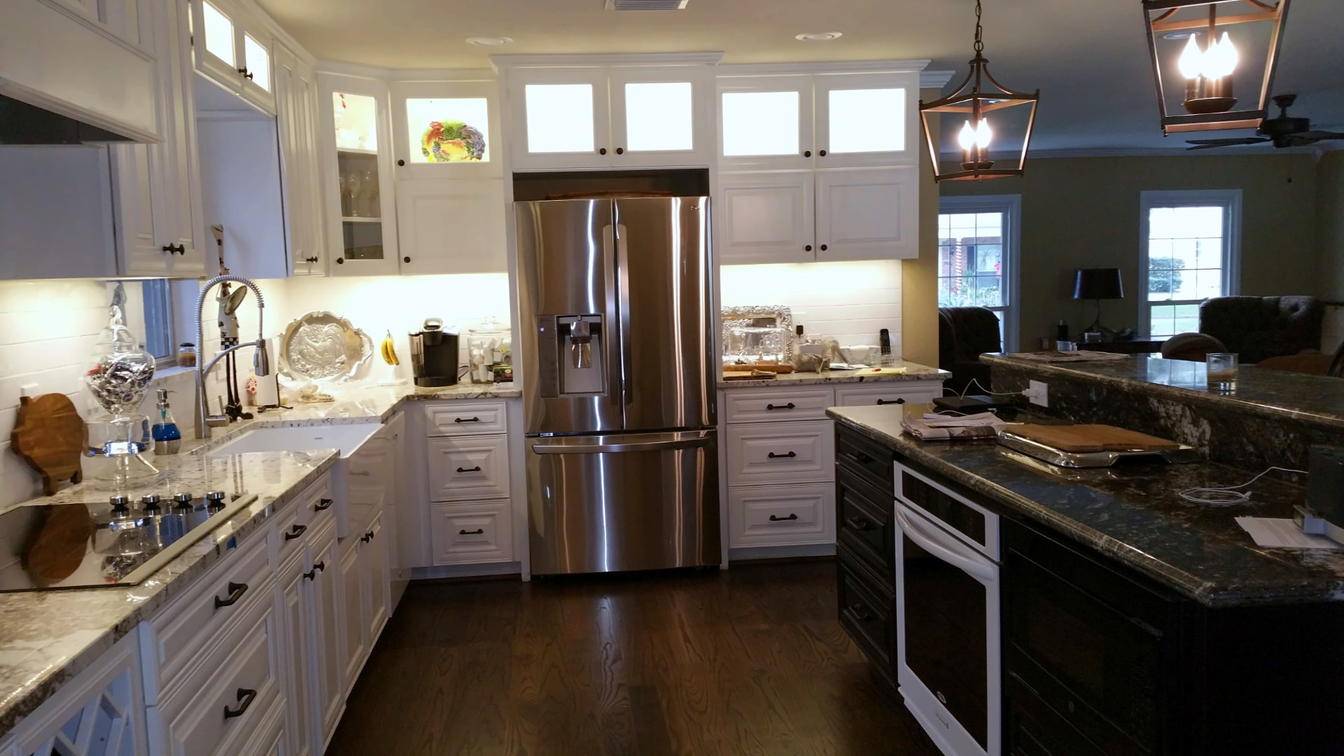 White paneled kitchen with a stainless refrigerator, lighted glass cabinets, and a dark island with black granite