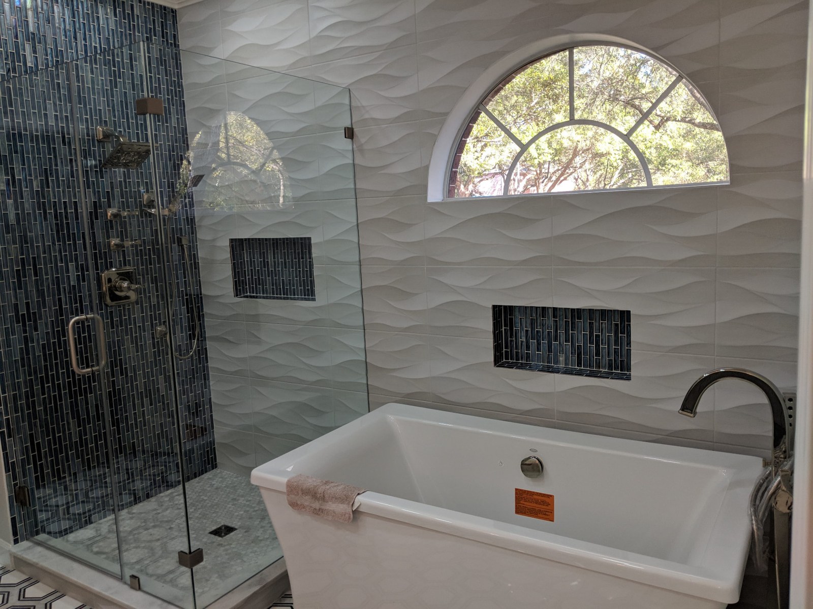Finished bathroom with blue mosaic shower tile, white sculpted wall tile, and a freestanding tub beneath an arched window