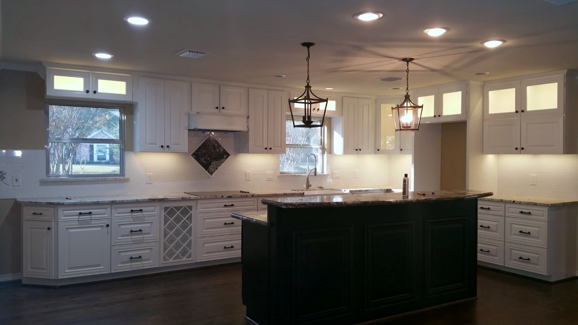 White kitchen with illuminated glass-front upper cabinets, a dark green island with granite top, and lantern pendant lights