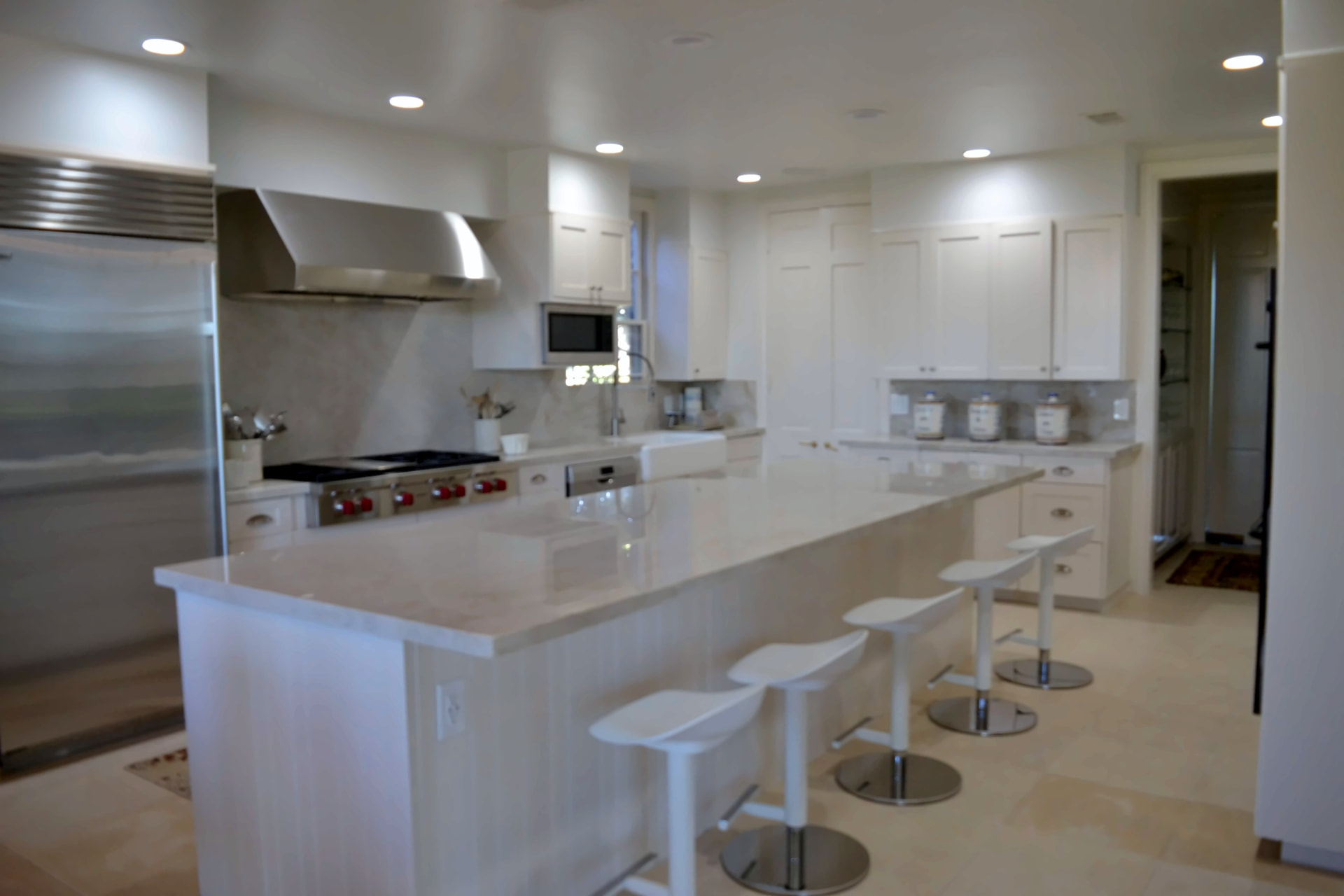 All-white luxury kitchen with a large marble island, white counter stools, professional range, and stainless hood